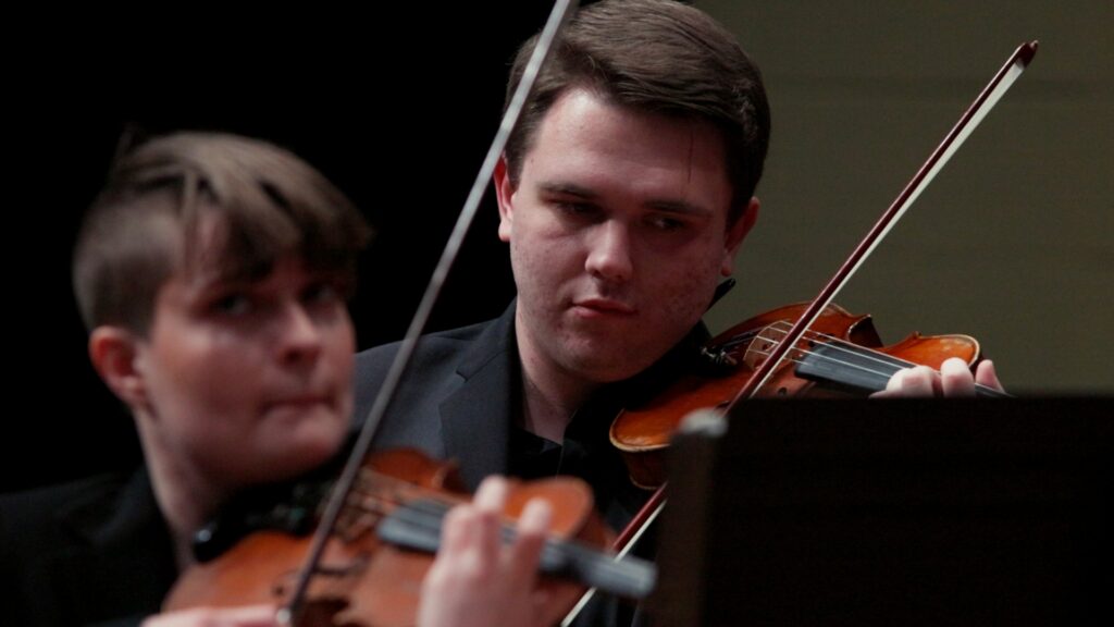 Photo of student musician playing the violin with another student violinist in the background.